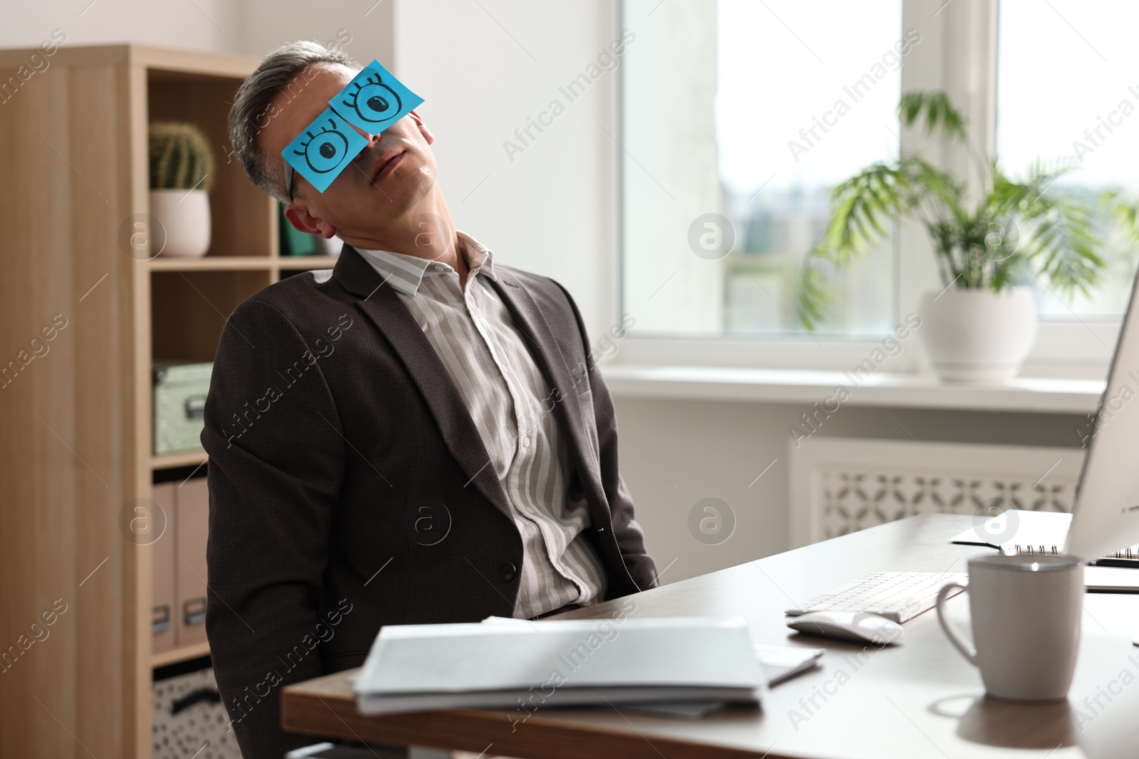 Photo of Man with fake eyes painted on sticky notes snoozing at workplace in office