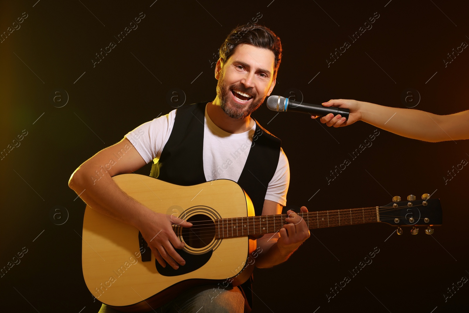 Photo of Handsome man with acoustic guitar singing while woman holding microphone on dark background, closeup