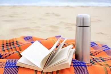 Metallic thermos with hot drink, open book and plaid on sandy beach near sea