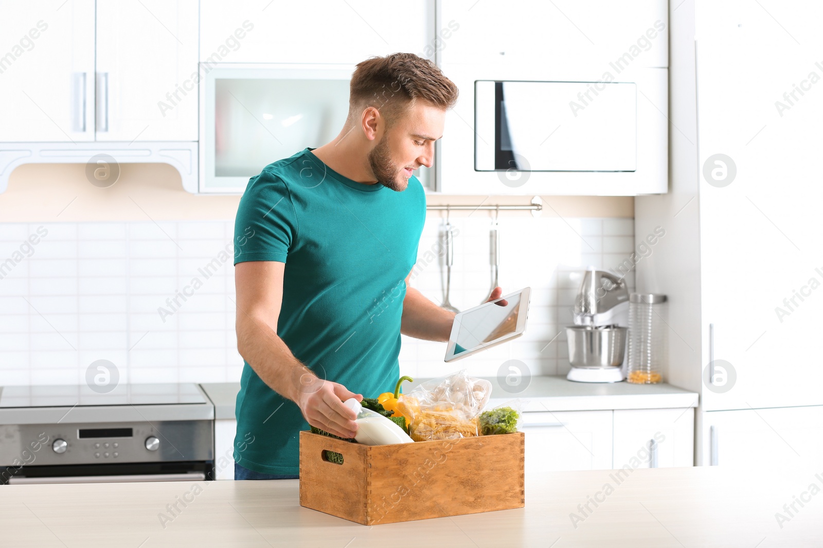 Photo of Man with wooden crate full of products and tablet in kitchen. Food delivery service