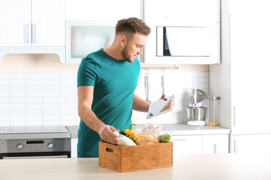 Photo of Man with wooden crate full of products and tablet in kitchen. Food delivery service