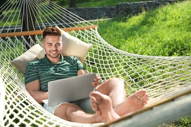 Young man with laptop resting in comfortable hammock at green garden