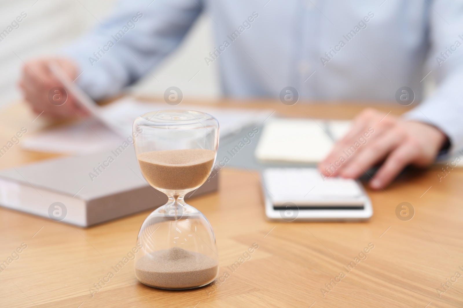 Photo of Hourglass with flowing sand on desk. Man using calculator indoors, selective focus