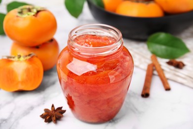 Jar of tasty persimmon jam and ingredients on white marble table