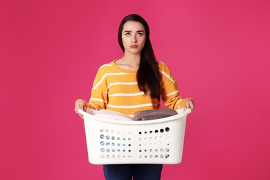 Displeased young woman holding basket with laundry on color background