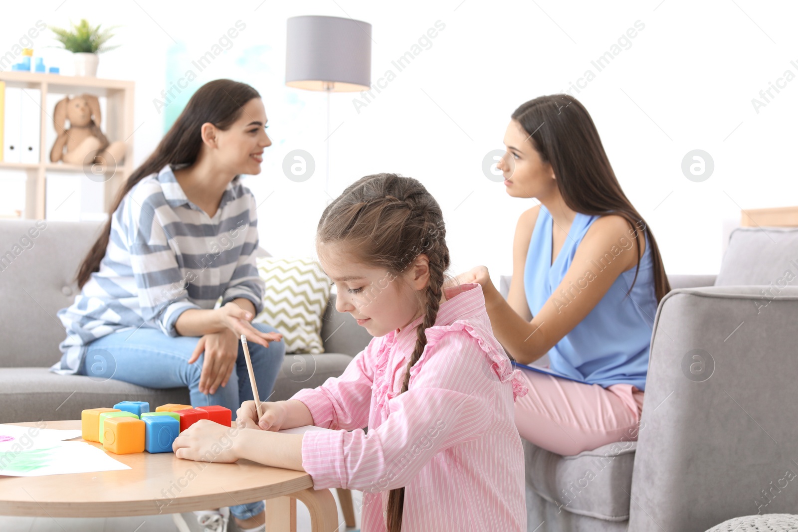 Photo of Little girl drawing at table while child psychologist talking with her mother in office