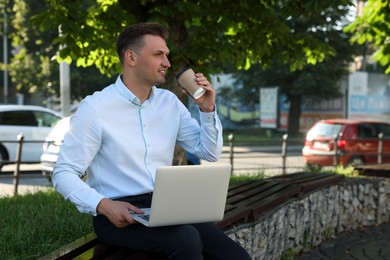 Photo of Handsome man with laptop drinking coffee on bench outdoors. Space for text