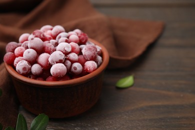 Photo of Frozen red cranberries in bowl and green leaves on wooden table, closeup. Space for text