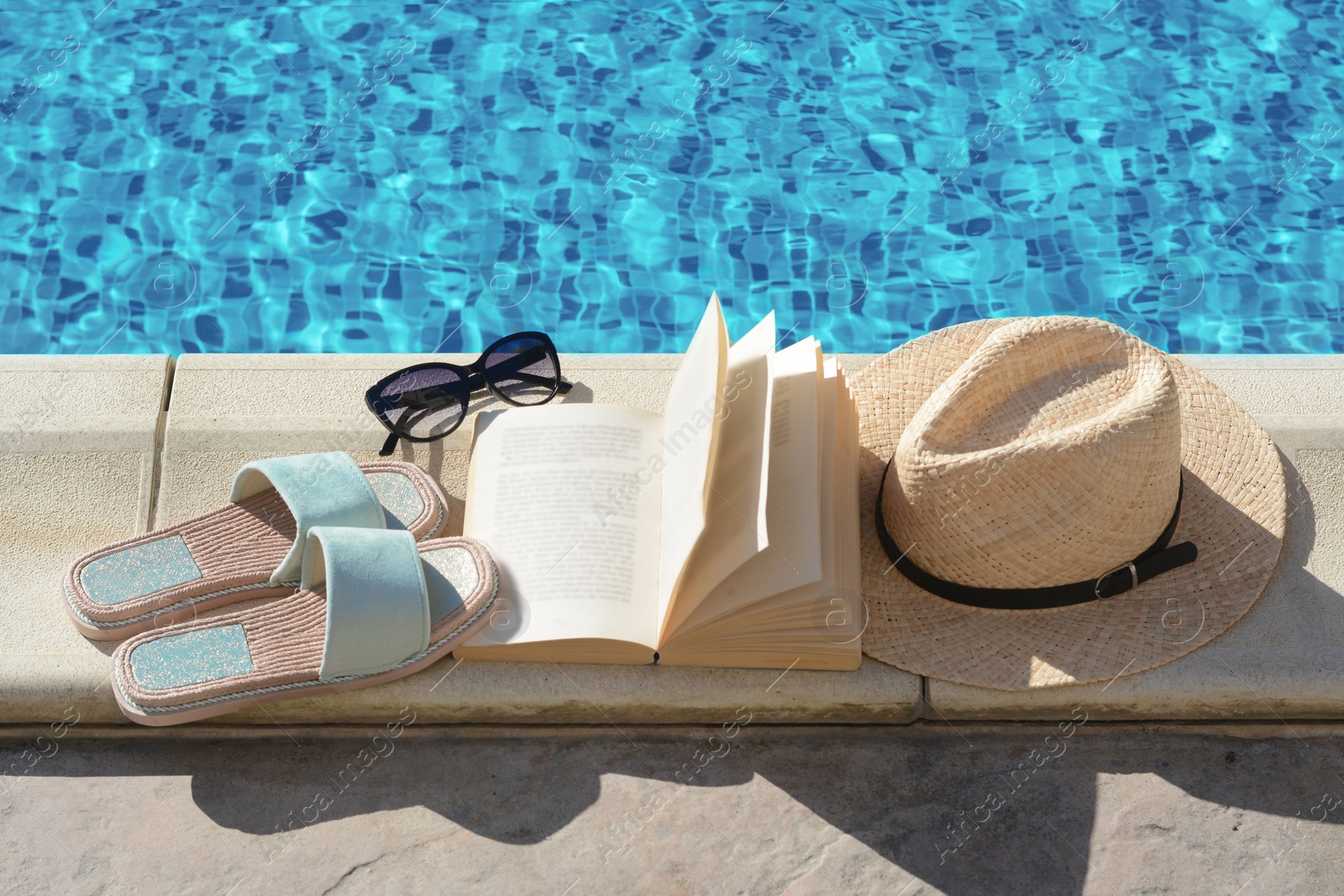 Photo of Stylish sunglasses, slippers, straw hat and book at poolside on sunny day. Beach accessories