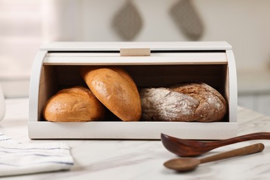 Photo of Wooden bread basket with freshly baked loaves and spoons on white marble table in kitchen