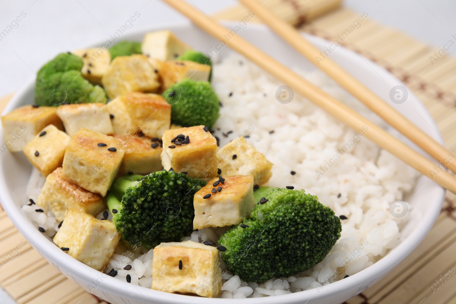 Photo of Bowl of rice with fried tofu and broccoli on bamboo mat, closeup