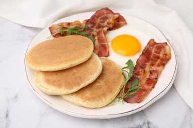 Photo of Plate with tasty pancakes, fried egg, arugula and bacon on white marble table, closeup