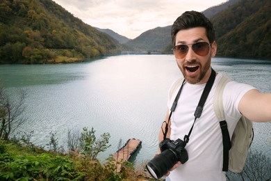 Image of Smiling man with camera taking selfie near beautiful river in mountains