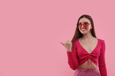 Photo of Pink look. Beautiful woman in heart shaped sunglasses pointing at something on color background, space for text