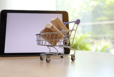 Photo of Internet shopping. Small cart with boxes near modern tablet on table indoors