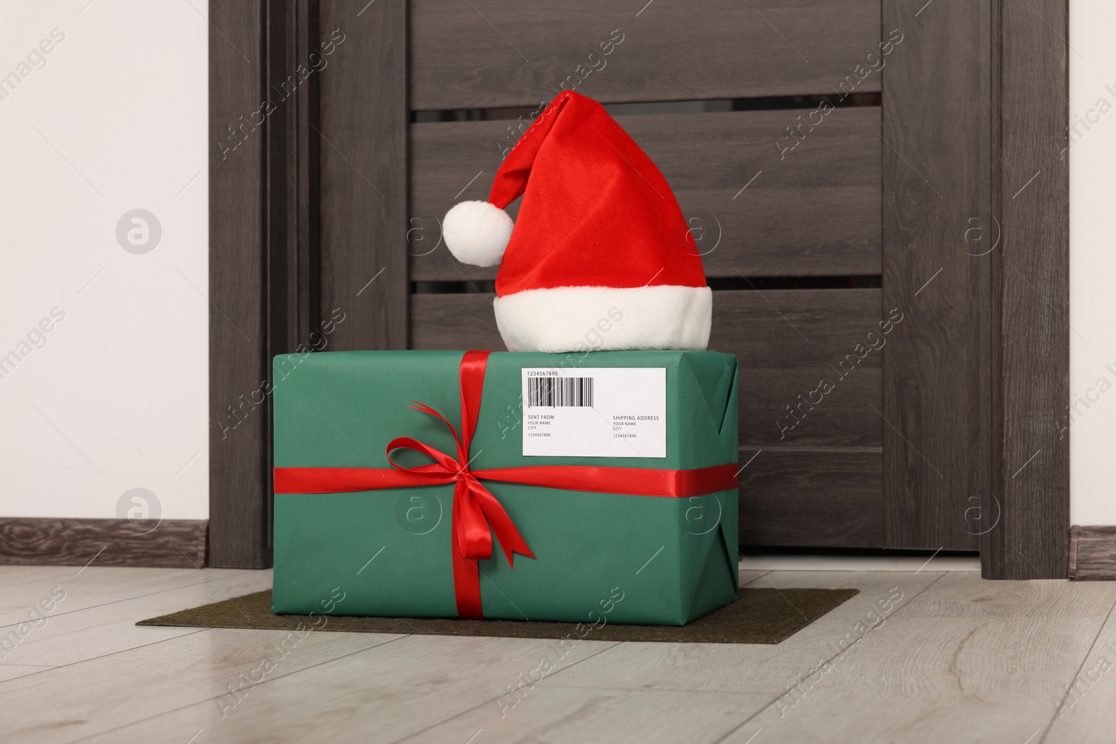 Photo of Christmas gift box and Santa hat near door indoors. Sending present by mail