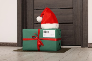 Christmas gift box and Santa hat near door indoors. Sending present by mail