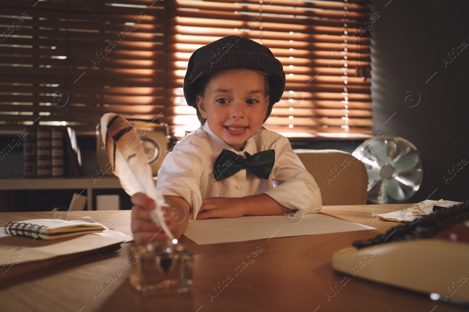 Photo of Cute little detective dipping feather into ink at table in office