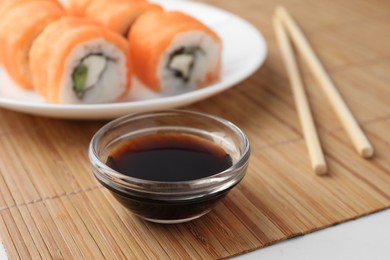 Photo of Bowl of tasty soy sauce, chopsticks and sushi rolls with salmon on bamboo mat, closeup