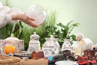 Photo of Woman holding glass vase above table with different stuff indoors, closeup. Garage sale