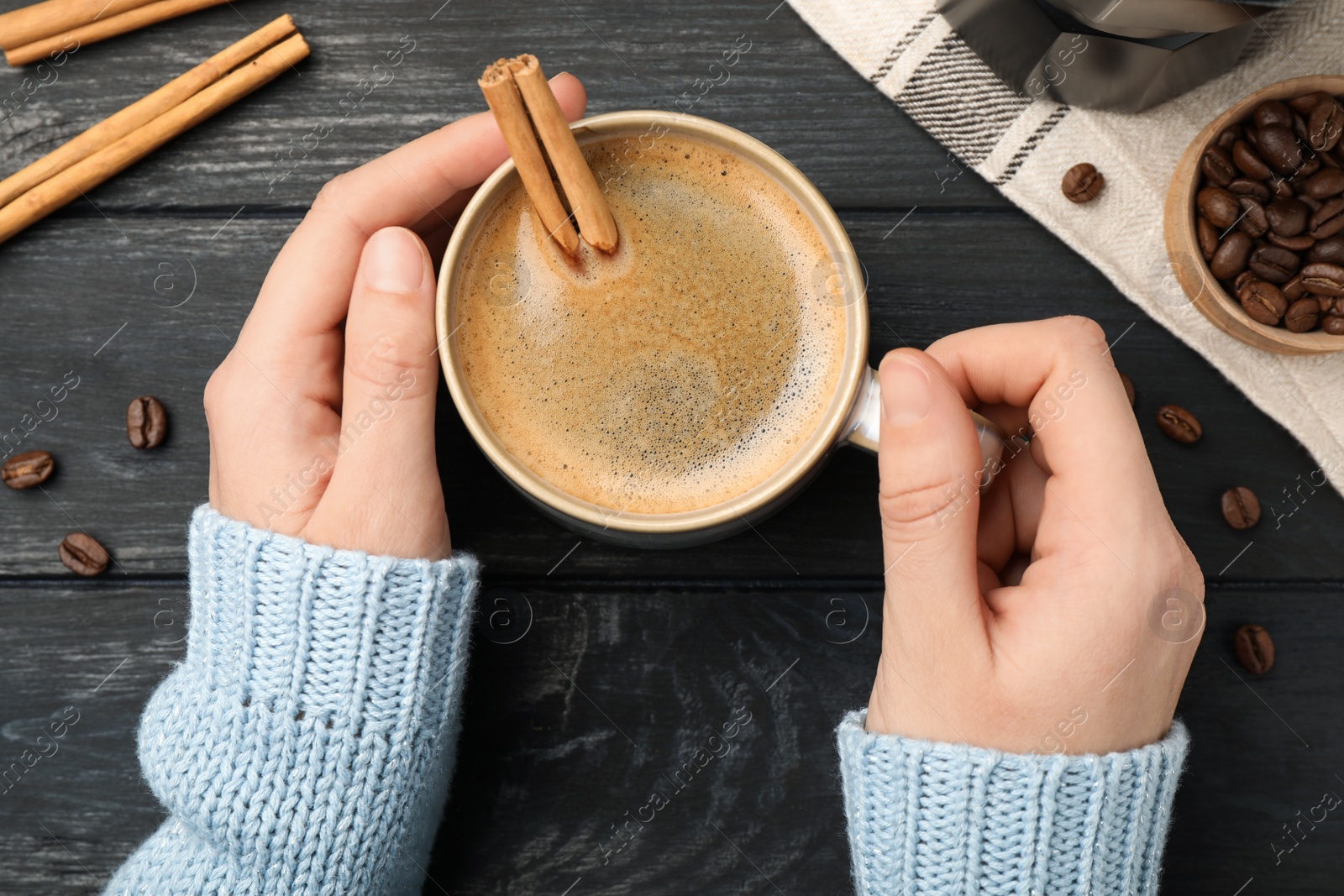 Photo of Woman holding cup of hot coffee with aromatic cinnamon at black wooden table, top view