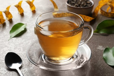 Cup of herbal diet tea, spoon, measuring tape and green leaves on brown marble table, closeup. Weight loss concept