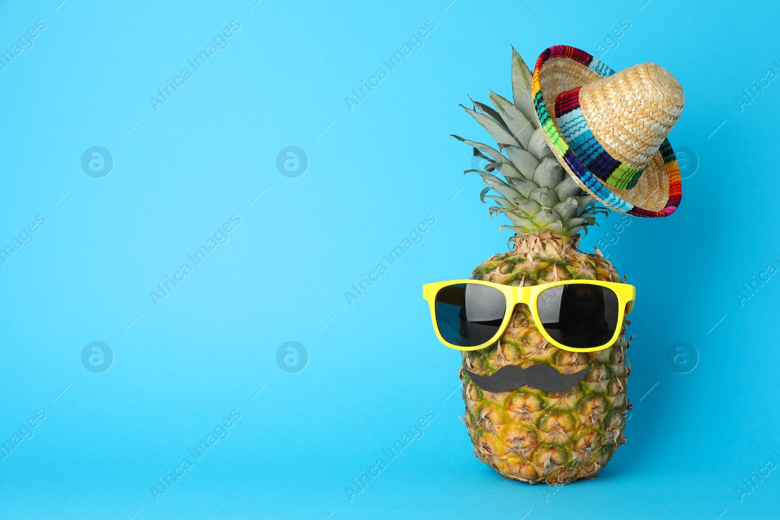 Photo of Pineapple with Mexican sombrero hat, sunglasses and fake mustache on light blue background, space for text