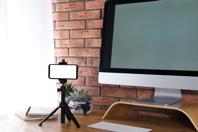 Photo of Tripod with smartphone near computer on wooden table indoors. Mockup for design