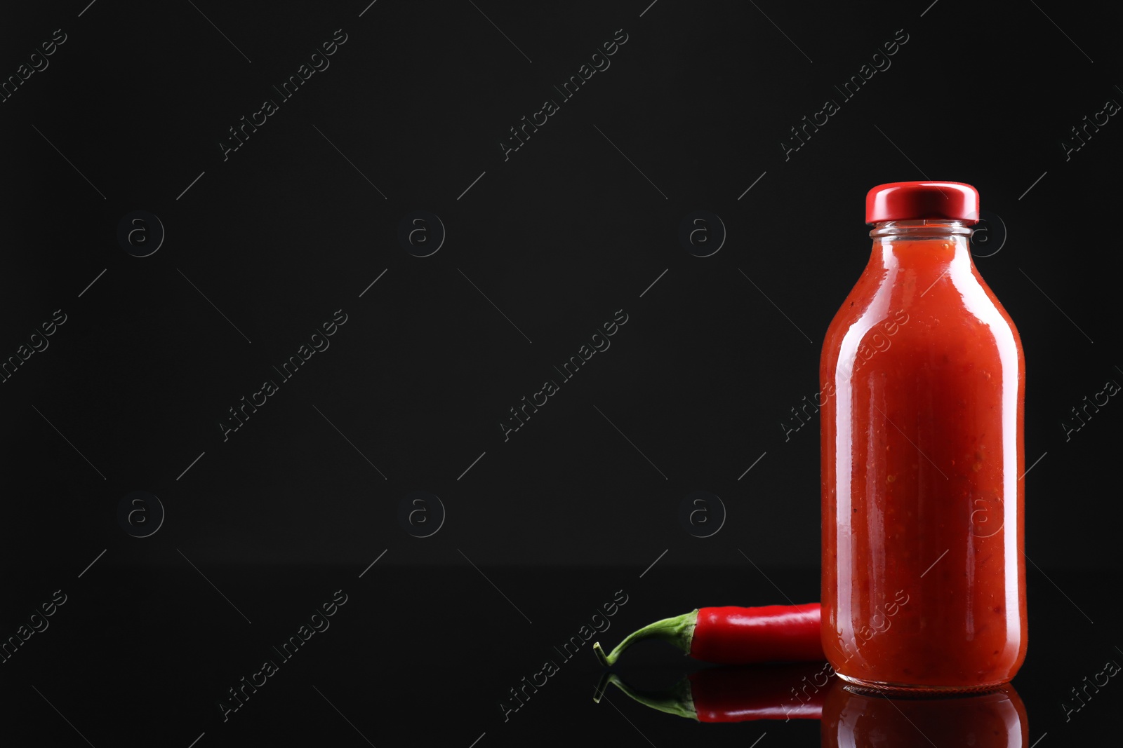 Photo of Spicy chili sauce in bottle and pepper against dark background, space for text