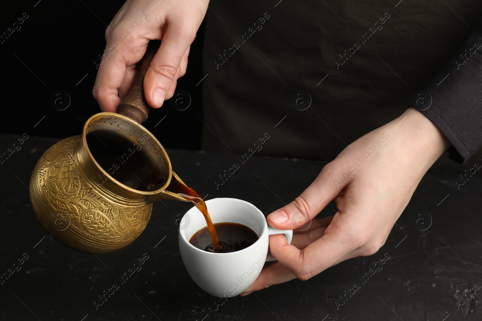 Photo of Turkish coffee. Woman pouring brewed beverage from cezve into cup at black table, closeup