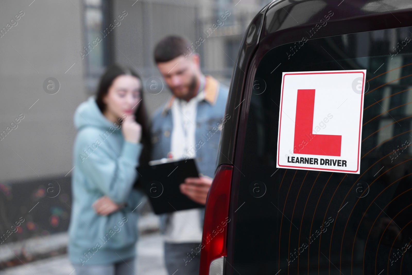 Photo of Learner driver and instructor with clipboard near car outdoors, selective focus on L-plate. Driving school