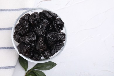 Photo of Sweet dried prunes in bowl and green leaves on white textured table, top view. Space for text