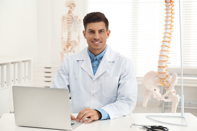 Photo of Male orthopedist with laptop near human spine model in office