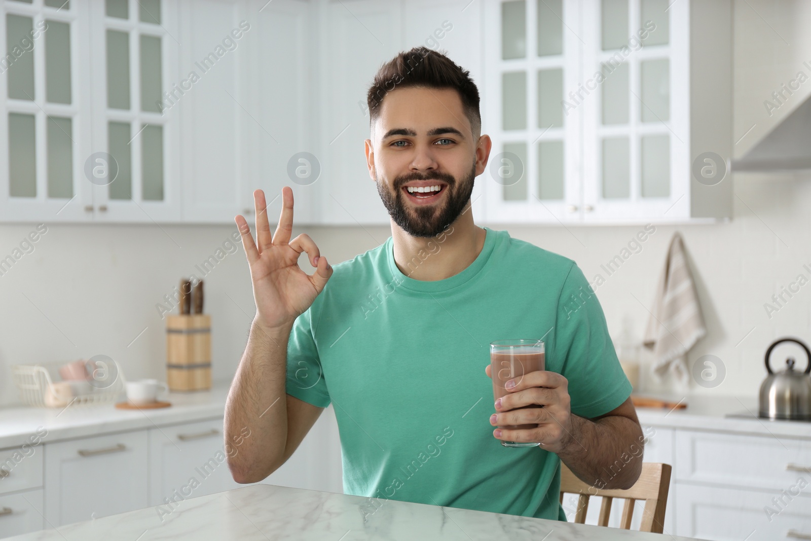 Photo of Young man with glass of chocolate milk showing Ok in kitchen