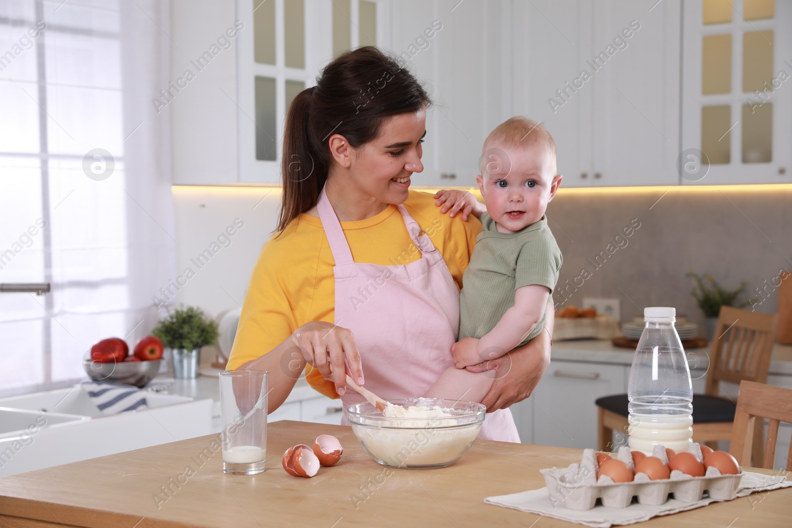 Photo of Happy young woman holding her cute little baby while making dough in kitchen