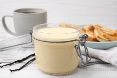 Photo of Tasty condensed milk in jar, crepes and vanilla pods on white wooden table, closeup