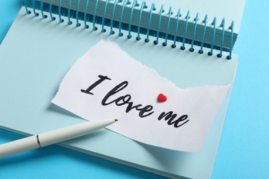 Photo of Paper with handwritten phrase I Love Me, notebook and pen on light blue background, closeup