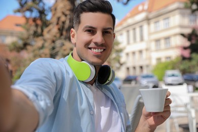 Photo of Happy man with cup of coffee and headphones taking selfie in outdoor cafe
