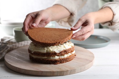 Photo of Woman stacking layers of sponge cake at white wooden table, closeup