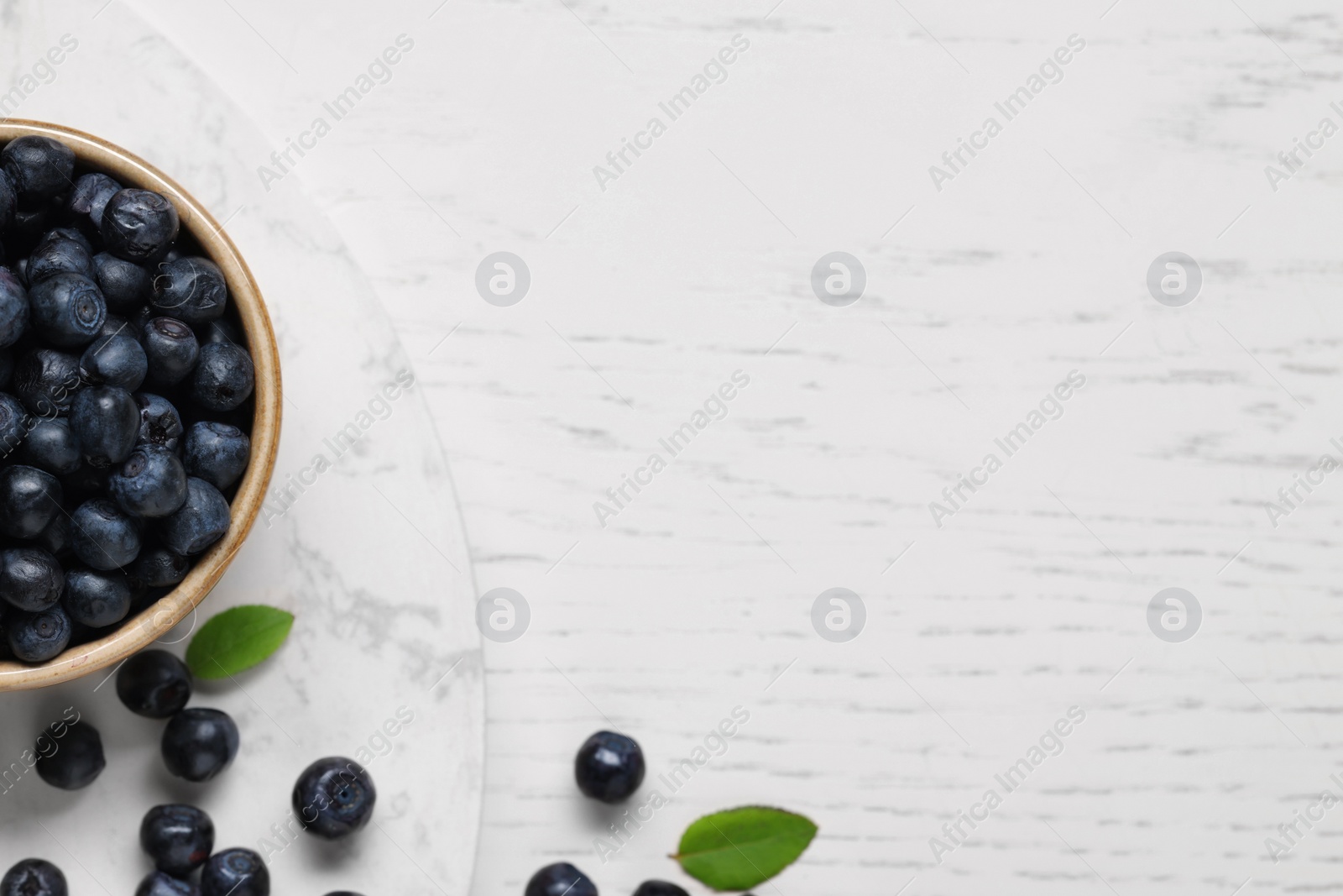 Photo of Bowl of tasty fresh bilberries and green leaves on white wooden table, flat lay. Space for text