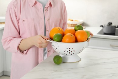 Photo of Woman holding colander with fresh fruits at white marble table in kitchen, closeup