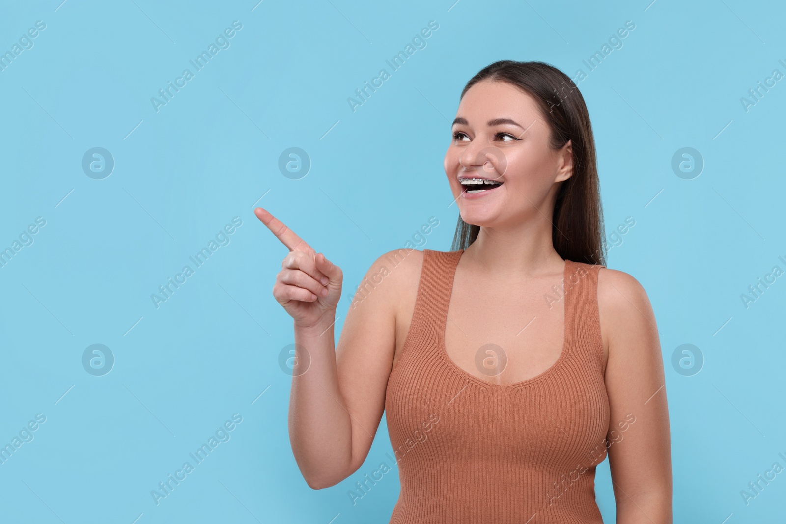 Photo of Smiling woman with dental braces pointing at something on light blue background. Space for text