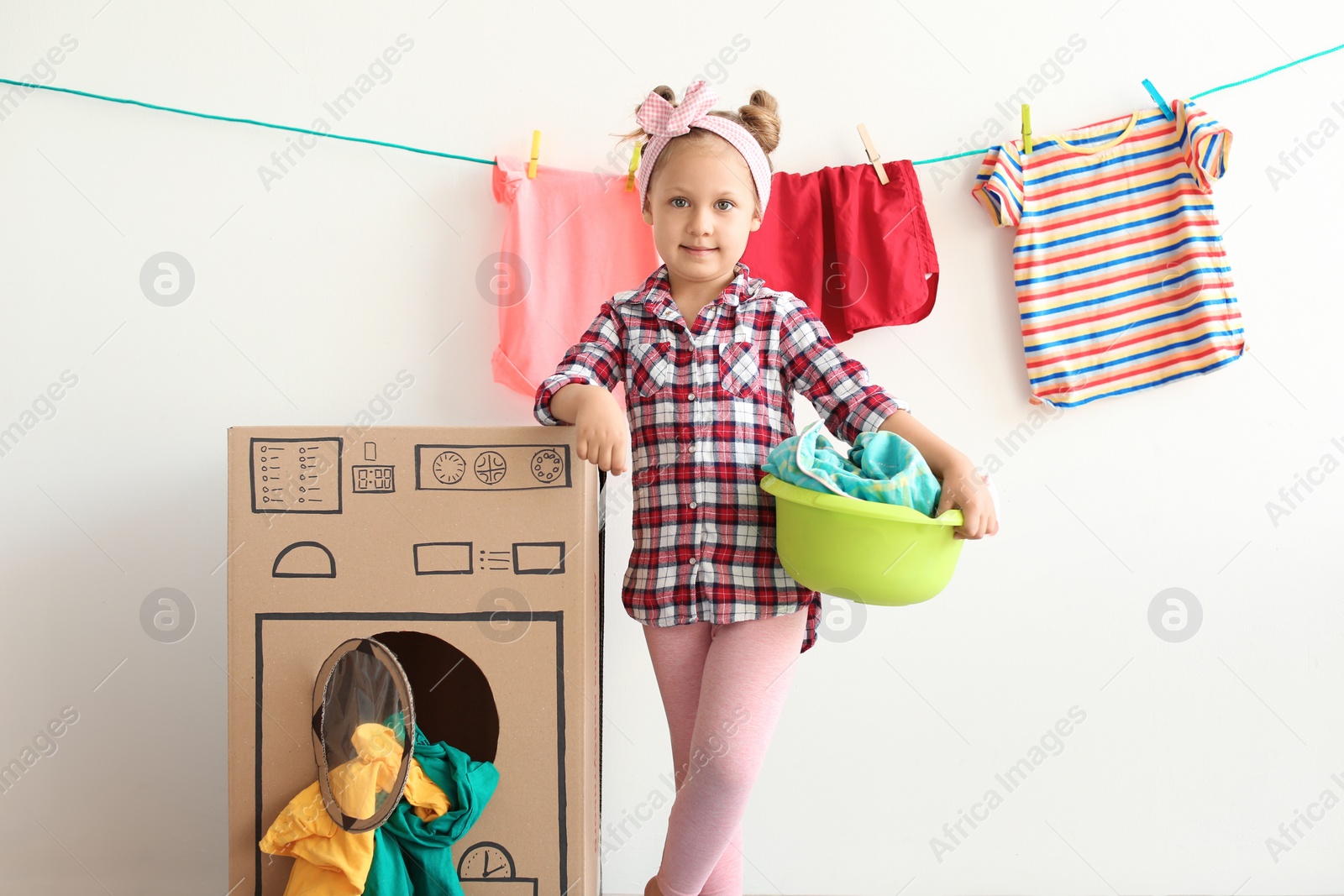 Photo of Adorable little child playing with cardboard washing machine and clothes indoors