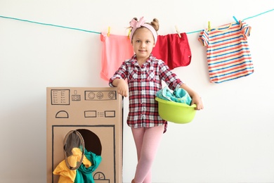 Adorable little child playing with cardboard washing machine and clothes indoors