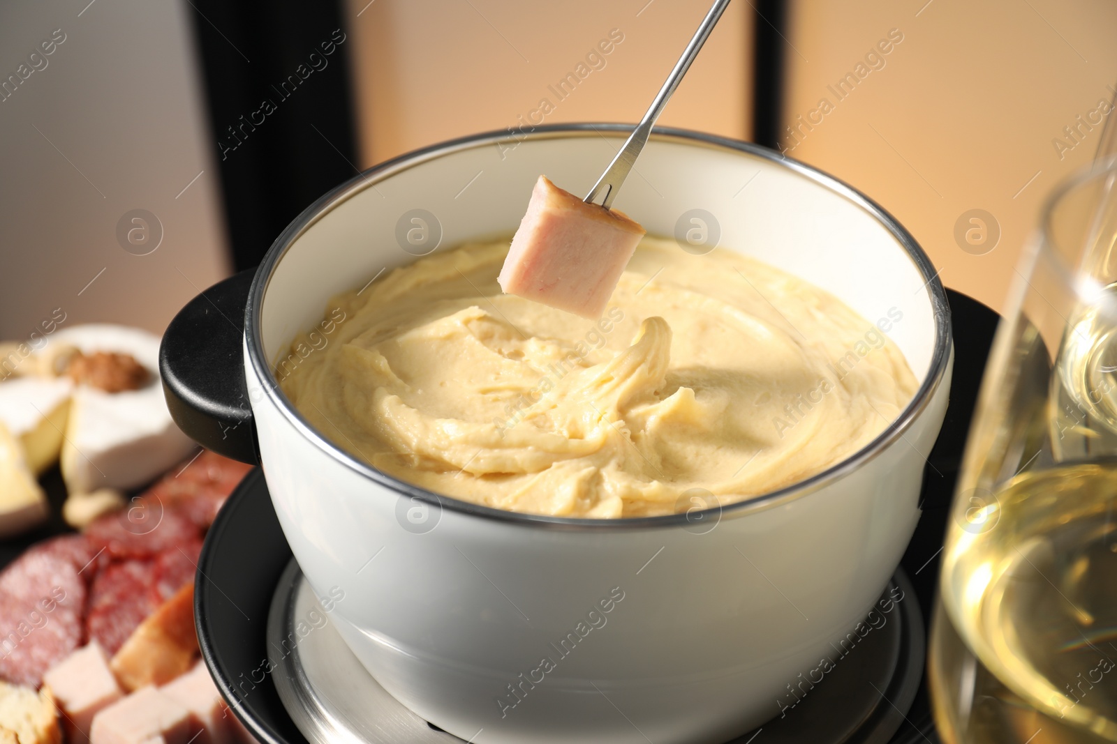 Photo of Dipping piece of ham into fondue pot with melted cheese at table with snacks, closeup