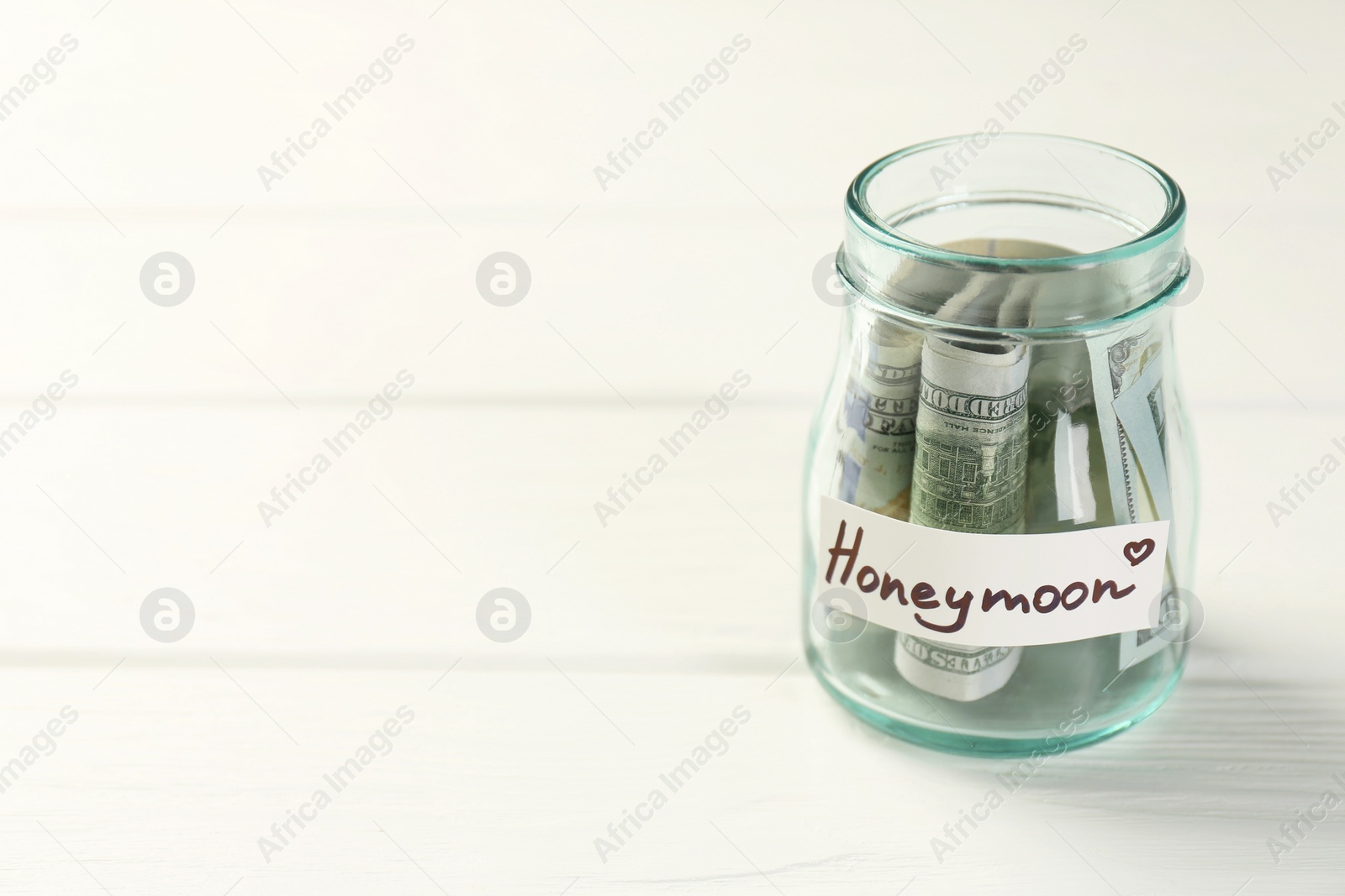 Photo of Glass jar with word Honeymoon and dollar banknotes on white wooden table, closeup. Space for text