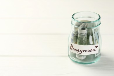 Glass jar with word Honeymoon and dollar banknotes on white wooden table, closeup. Space for text