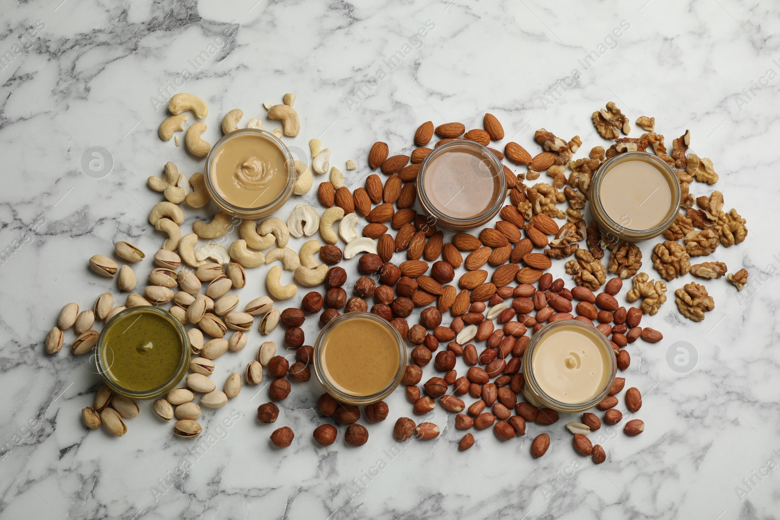 Photo of Jars with butters made of different nuts and ingredients on white marble table, flat lay