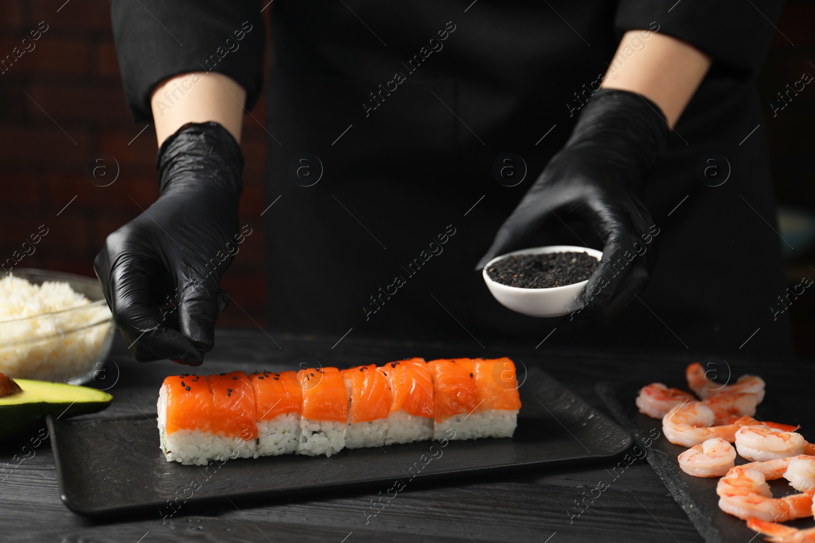 Photo of Chef in gloves adding sesame seeds onto tasty sushi rolls at black wooden table, closeup