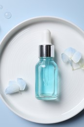 Bottle of cosmetic serum and beautiful flowers on light blue background, top view